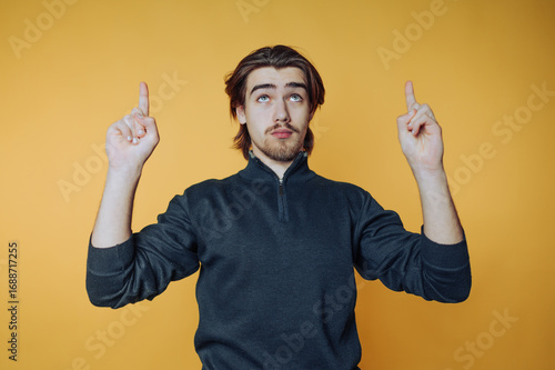 portrait of young man dressed in a dark turtleneck is pointing upwards with a contemplative look on yellow background