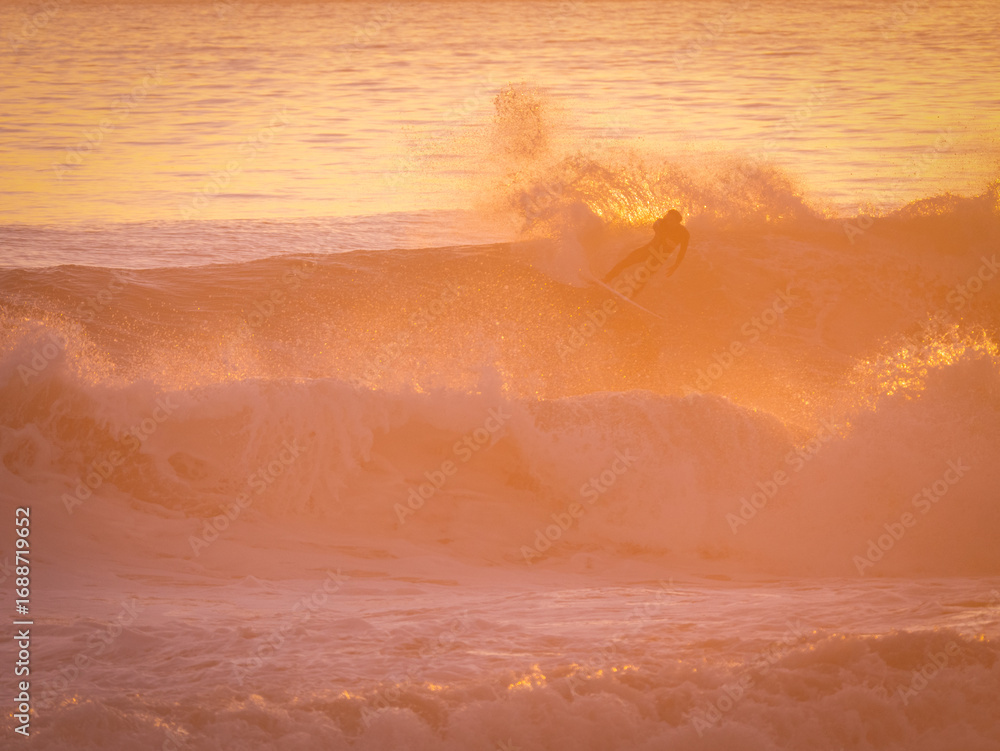 Fototapeta premium SILHOUETTE: Unknown surfer carves along the face of a breaking wave, backlit by intense golden light of setting sun. As he makes a top turn, water sprays around. Riding waves at famous Moroccan spot.