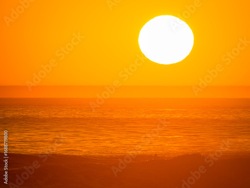 LENS FLARE, CLOSE UP: Sun hangs over the ocean horizon, casting an intense warm orange glow. Water and sky are a monochromatic study in gold, emphasizing dramatic beauty of evening light in Morocco.