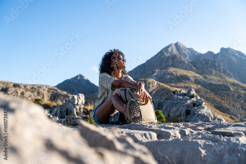 Bild auf Leinwand Young female traveler sitting on a rock with her backpack, enjoying a peaceful break in the Serra de Tramuntana mountains, Majorca