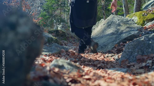 Tripod-level shot of a hiker walking down a rocky, leaf-covered mountain trail past the camera, with blurred trees in the background.