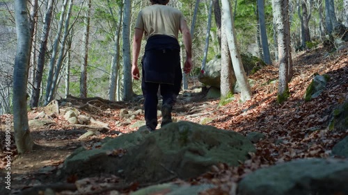 Tripod-level shot of a full-body hiker walking uphill on a rocky forest trail surrounded by trees, capturing natural movement and scenery.