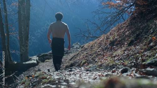 Tripod shot of a hiker walking along a leaf-covered mountain trail leading to a rocky and grassy clearing, with forested mountains in the background.