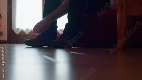 Tripod shot of a man tying mountain boots in backlight, showing legs and feet, then walking out of frame.