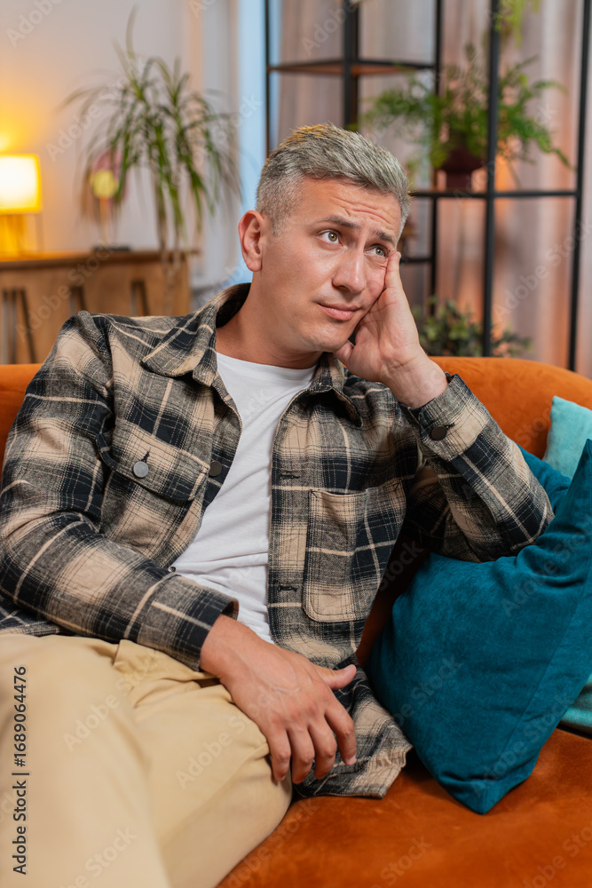 Obraz premium Thoughtful Caucasian man guy male sitting at home looks pensive thinks over life concerns or unrequited love, suffers from unfair situation. Problem, break up, depressed feeling bad annoyed, burnout.