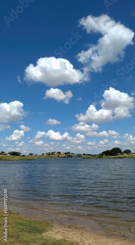 Serene waterscape of a lake under a bright blue sky dotted with puffy white clouds, bordered by grassy land and distant trees on the horizon