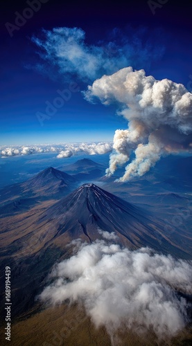 Aerial view of a dark, cone-shaped mountain with smoke billowing against a vibrant blue sky dotted with fluffy white clouds, surrounded by hazy landscapes
