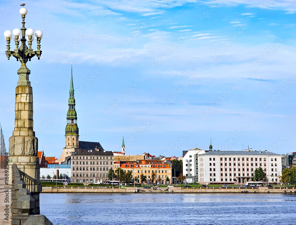 Fototapeta premium Panorama of the Riga Old Town with the spire of St. Peter's Church on the bank of the Daugava River and a bridge across the river with old lanterns.