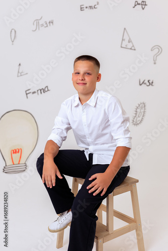 Thoughtful schoolboy in uniform sitting on wooden stool, student portrait with science and math formulas on white background, education and learning concept