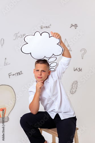 Thoughtful schoolboy in uniform sitting on wooden stool, student portrait with science and math formulas on white background, education and learning concept