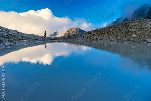 A person stands on rocky terrain, gazing at a stunning turquoise mountain lake surrounded. Dressed in hiking gear and holding trekking poles. Wildsee Pizol, Switzerland.