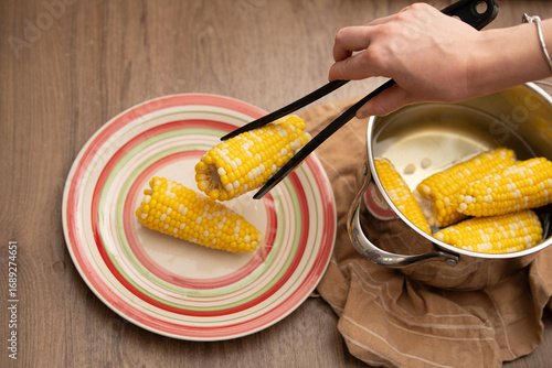 Fotografie A hand uses tongs to transfer steaming corn on the cob from a pot to a vibrant plate