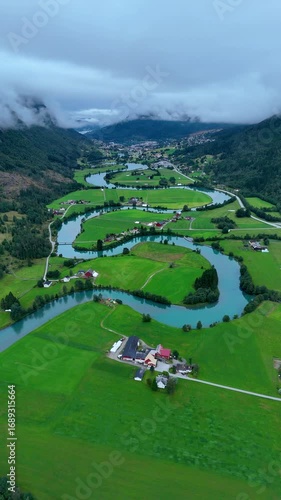 Majestic landscape of Stryn, Norway, reveals a winding river embraced by vibrant green fields and tranquil farms. The atmosphere is serene, with misty mountains in the background.