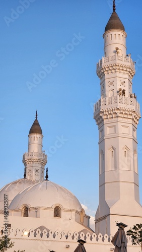 Quba Mosque, located in Medina, Saudi Arabia, is highlighted against a clear blue sky during sunset. The mosque features a prominent white facade with two minarets on either side.