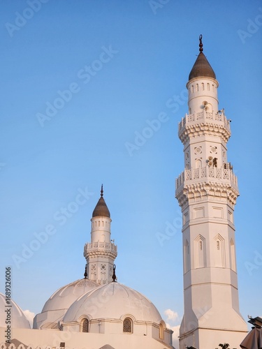 Quba Mosque, located in Medina, Saudi Arabia, is highlighted against a clear blue sky during sunset. The mosque features a prominent white facade with two minarets on either side.