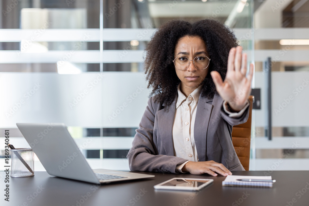 Obraz premium Portrait of an African American woman sitting at a desk in an office and showing a stop and warning sign to the camera with her hand