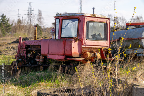 Old red crawler tractor with cabin, rusty and abandoned, standing among grass and bushes in a rural field.
