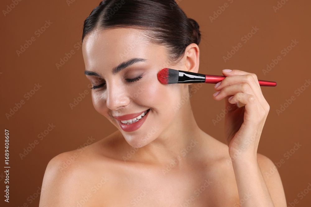 Fototapeta premium Beautiful woman applying makeup with brush on brown background