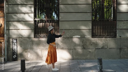 Chinese tourist smiling while vlogging on Madrid street with smartphone. Concept of travel influencer lifestyle, cultural exploration, and digital tourism
