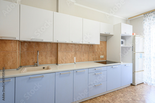 bright kitchen featuring white upper cabinets, blue lower cabinets, and a tiled backsplash. A sink and refrigerator are visible