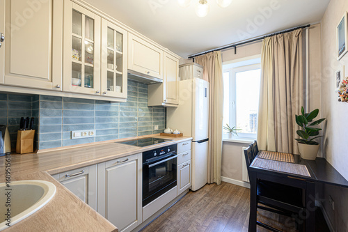 bright kitchen features beige cabinetry, blue tiled backsplash, and wooden countertops. black oven is built into cabinets, next to white refrigerator. black table and chairs are near window