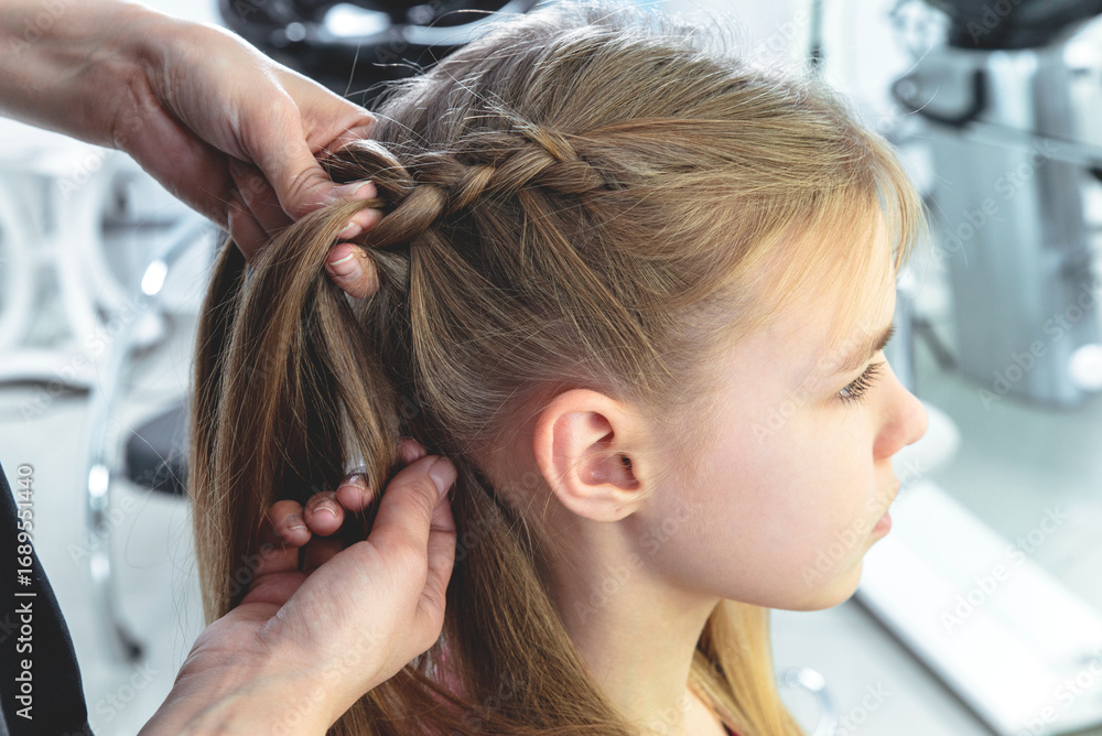 Fototapeta premium Hairdresser weaves a braid to a preteen blond girl in a beauty and hair salon