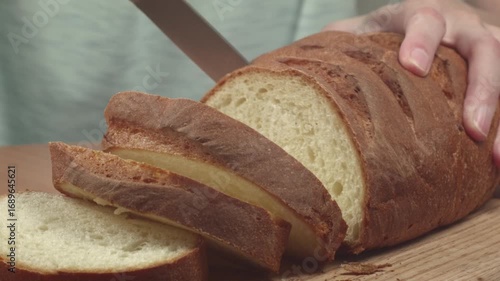 Close-up of young woman slicing fresh white bread with a kitchen knife on a cutting board in her kitchen, recorded with ambient sound.