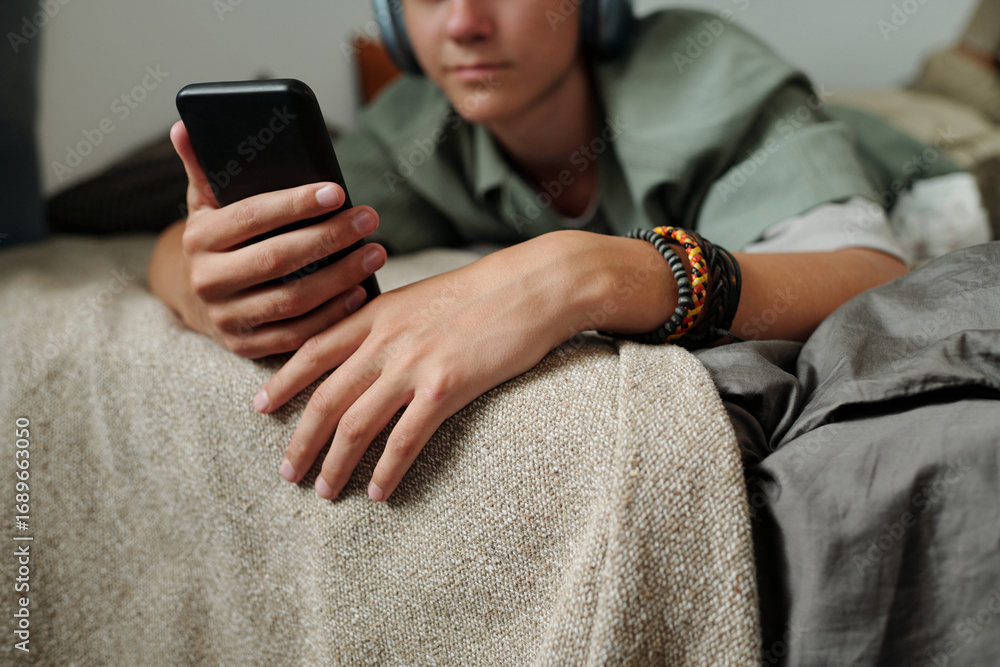 Fototapeta premium Teenager boy with light skin lying on bed using smartphone wearing headphones focusing on screen showing signs of gadget addiction with relaxed posture