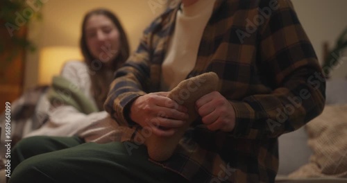 Close up of guy giving foot massage to his girlfriend who is sitting on sofa in socks during cozy romantic evening at home. Showing tenderness and care in massage