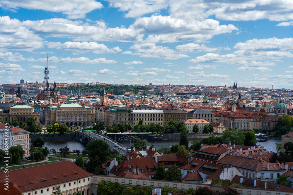 Fototapeta premium Panoramic cityscape featuring a river running through a historic European city under a bright blue sky with fluffy white clouds.