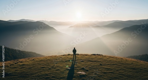 Solitary figure on grassy hilltop overlooking misty mountain valley at sunrise