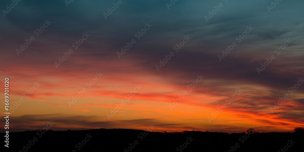 Fototapeta premium A fiery sunset paints the sky with vibrant colors over a silhouetted landscape.
