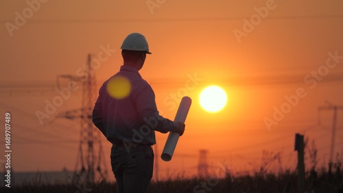 Power engineer working on power transmission line supports repair, sunset. Engineer man working on high voltage power plant construction project. Civil engineer reading drawing. Solar energy, nature