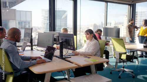 Group of colleagues working at computer stations in a bright modern office with city views, combining focus and collaboration while analyzing data and discussing tasks in a shared workspace.