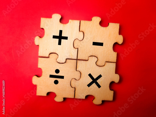The image shows four wooden puzzle pieces that together form a square. Each piece features a different basic arithmetic operation symbol