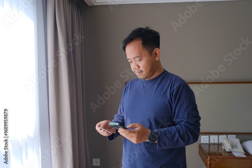A man in a navy long-sleeve shirt stands near a window, holding a smartphone and a credit card in his hands. The room is softly lit with natural light, creating a calm indoor atmosphere.  
