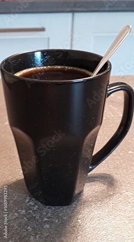 A dark, steaming liquid fills a matte black mug with a spoon resting inside, sitting on a neutral-colored surface with cabinetry in the background