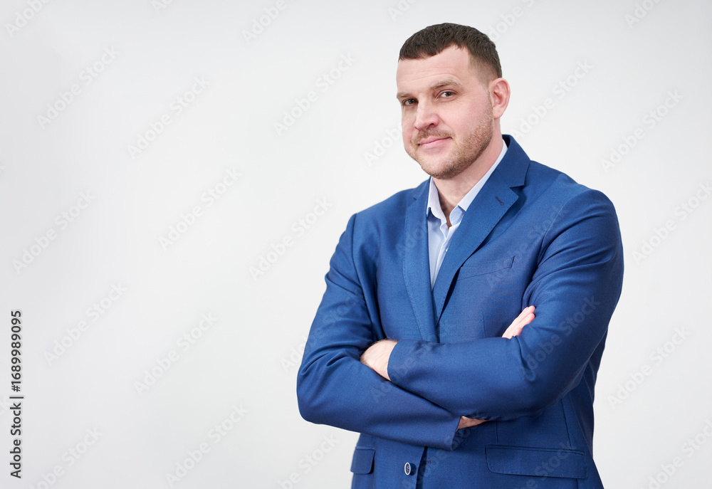 Successful young businessman with crossed arms wearing blue suit with a lovely genuine smile posing against grey background