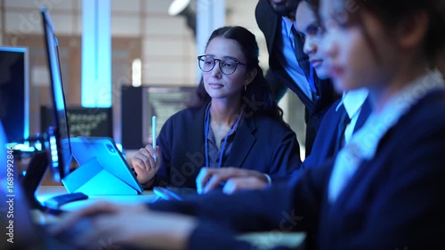 A team of young professionals works together late at night in a modern office. They are looking at a tablet and computer screens, discussing a new and important project.