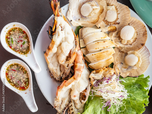 Grilled seafood platter with river prawns, squid, and scallops served with spicy Thai seafood dipping sauce and fresh vegetables on white plate.