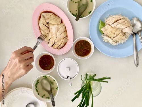 A woman is scooping a plate of Betong chicken, sliced and paired with steamed rice, drizzled with special sauce and green onions, accompanied by a chicken bone soup on a white table
