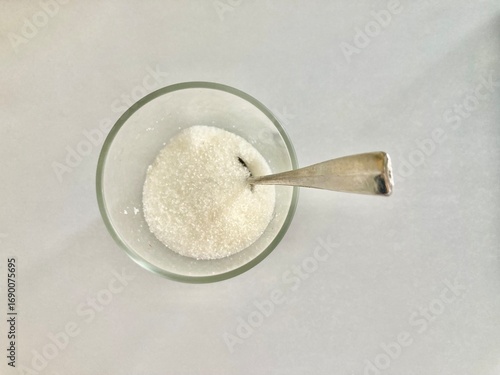 top view shot of a clear glass cup of white sugar and a silver spoon put on a white table