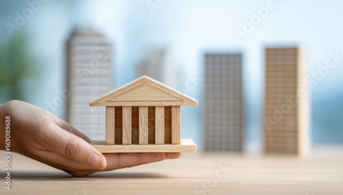 A hand holds a small wooden model of a building, with city models in the background