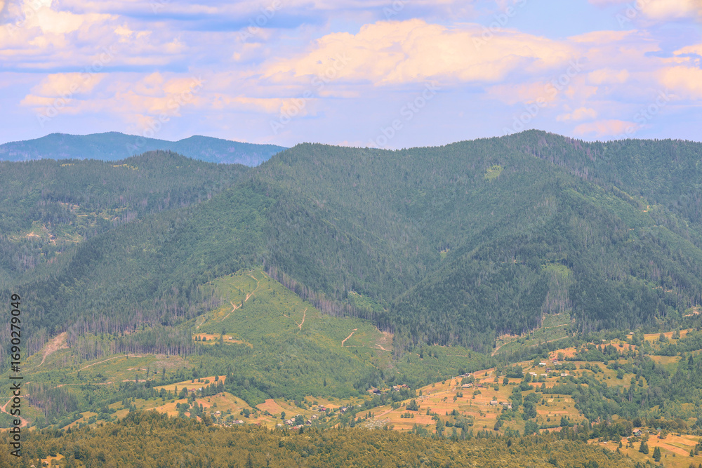 Fototapeta premium Endless mountain slopes covered with forest open up a fascinating view of a cozy village in the valley and fields. The warm sunlight and the colors of the sky create a sense of peace and idyll.