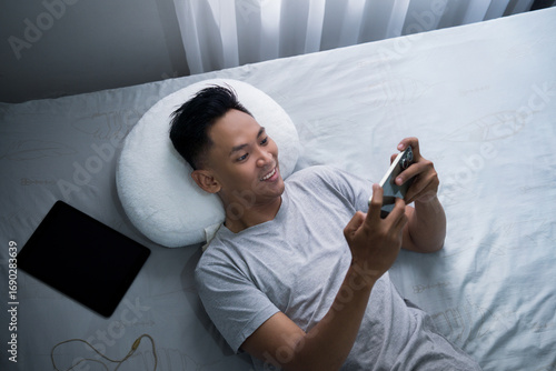 Asian young man enjoys leisure time playing mobile game in bed on his smartphone, surrounded by personal items such as headphones and a tablet.