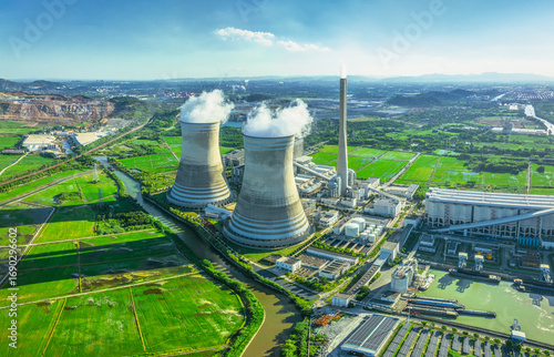 Aerial view of a large thermal power station with cooling towers emitting steam, situated in a green rural landscape with farmland.