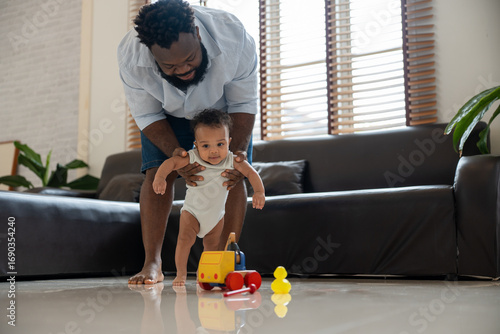 Quadro em tela Happy African American dad holding adorable infant son helps him take first steps walking in living room at home