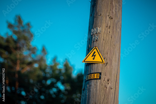 A wooden utility pole displays a yellow high voltage warning sign, with a vibrant blue sky and trees in the background.
