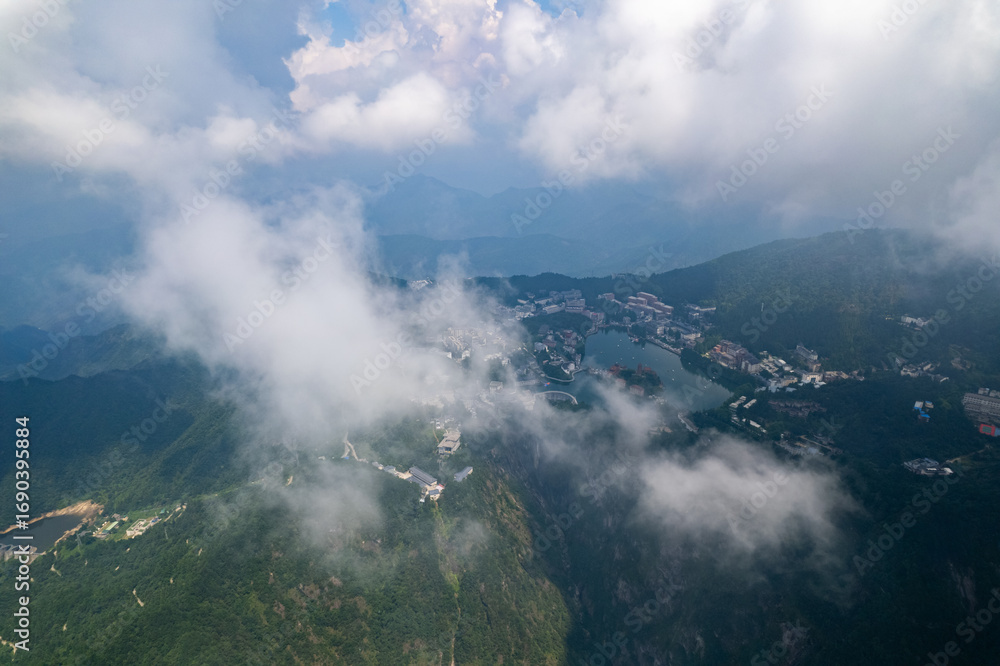 Fototapeta premium Jiugong Mountain in Xianning City China unfolds below a blanket of soft clouds, revealing a serene vista of lush green slopes and a tranquil lake surrounded by buildings.