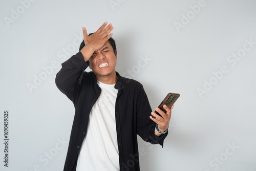 Asian man holding smartphone and slapping forehead with regret expression. Concept of mistake, failure, disappointment, and stress reaction on white background.